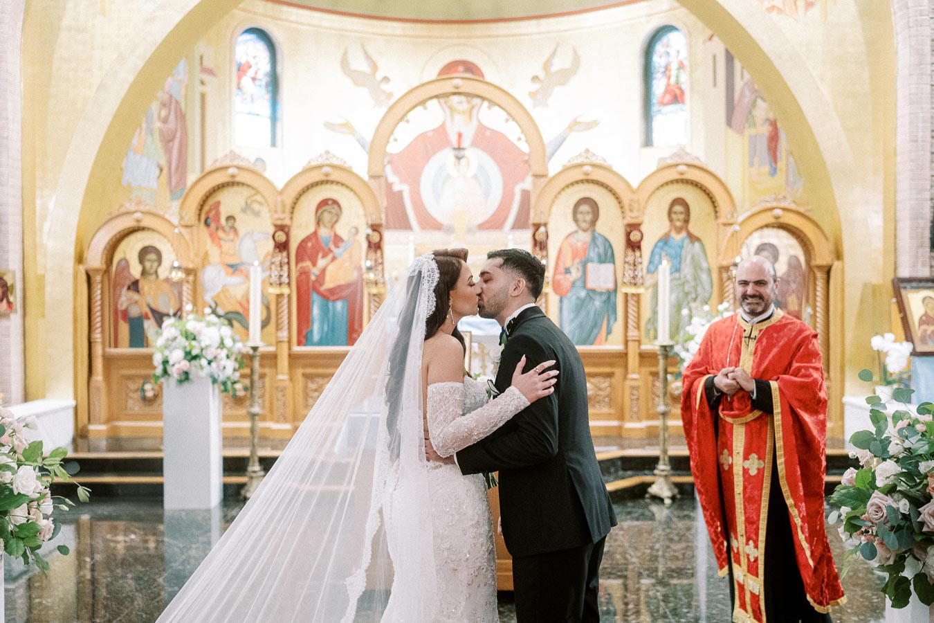 A bride and groom share a kiss in a beautiful ornate church during their wedding ceremony, with a priest smiling in the