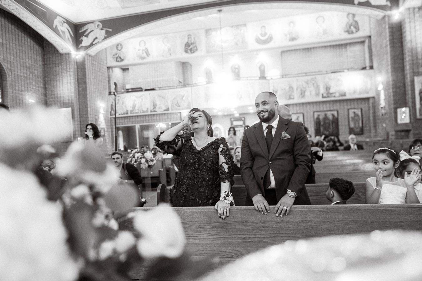 Black and white image of a joyful moment in a church during a wedding ceremony. A woman in a lace dress and a man in a suit,