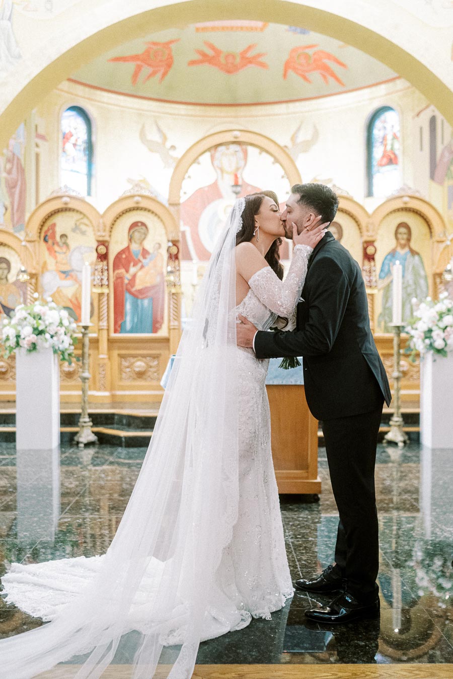 Bride and groom share a romantic kiss at the altar of an ornate church, surrounded by floral arrangements and religious