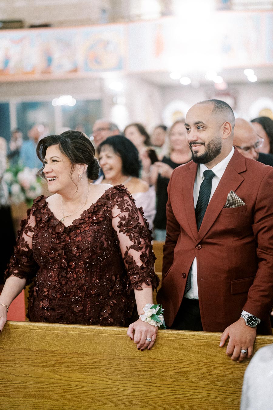Two people smiling at a joyful ceremony inside a decorated venue. The woman is wearing an elegant maroon dress with floral