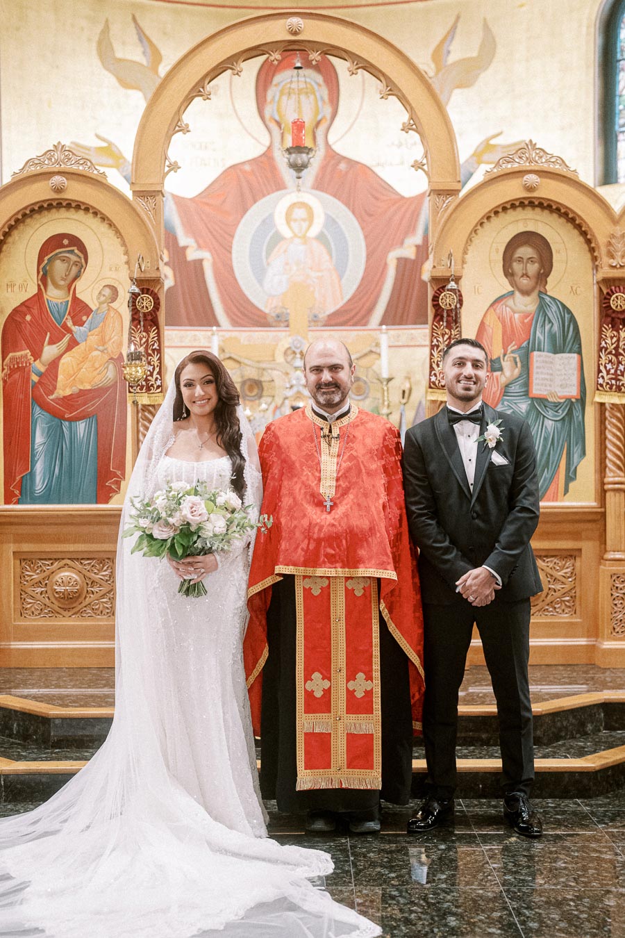 A bride and groom stand with a priest in an ornate church adorned with religious icons, celebrating a Christian wedding