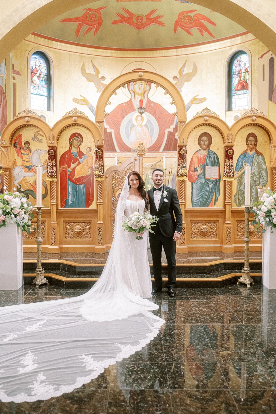 A bride and groom pose in a traditional church setting, surrounded by ornate religious icons and floral arrangements. The