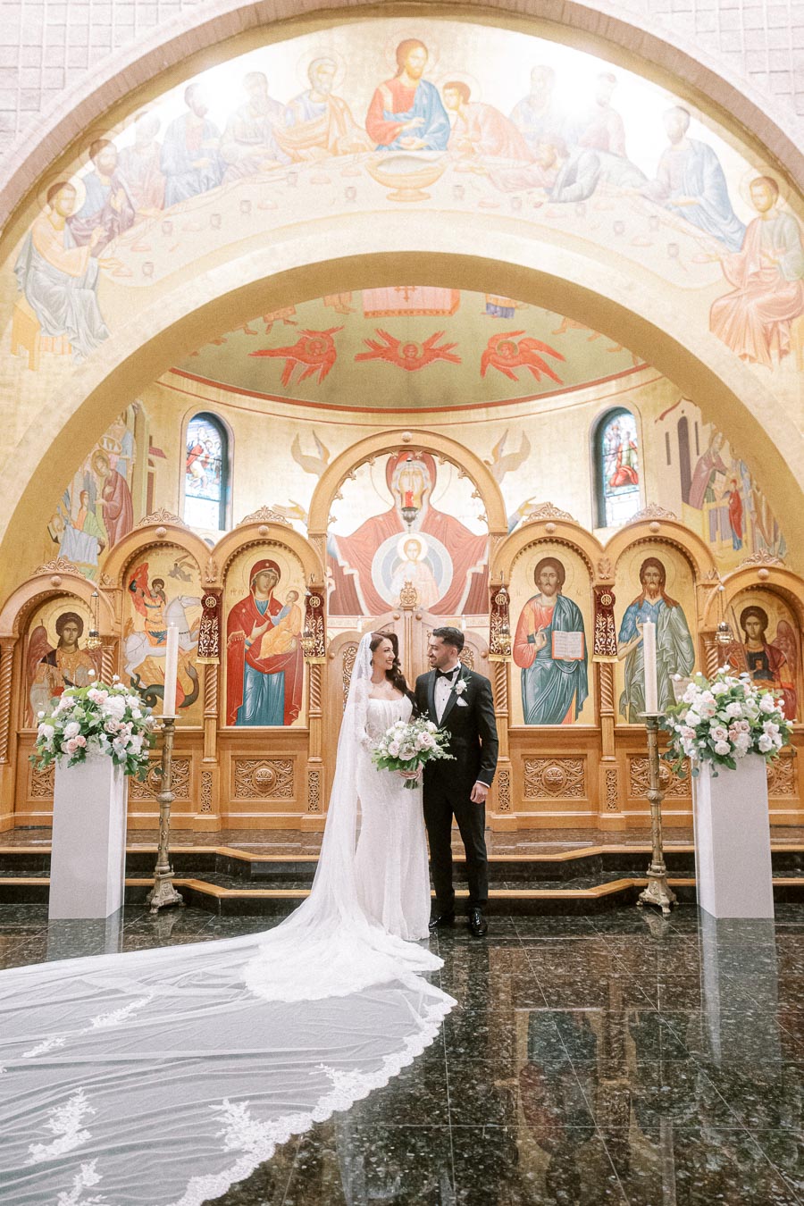 Bride and groom standing under a dome in an ornately decorated church with religious icons in the background, surrounded by
