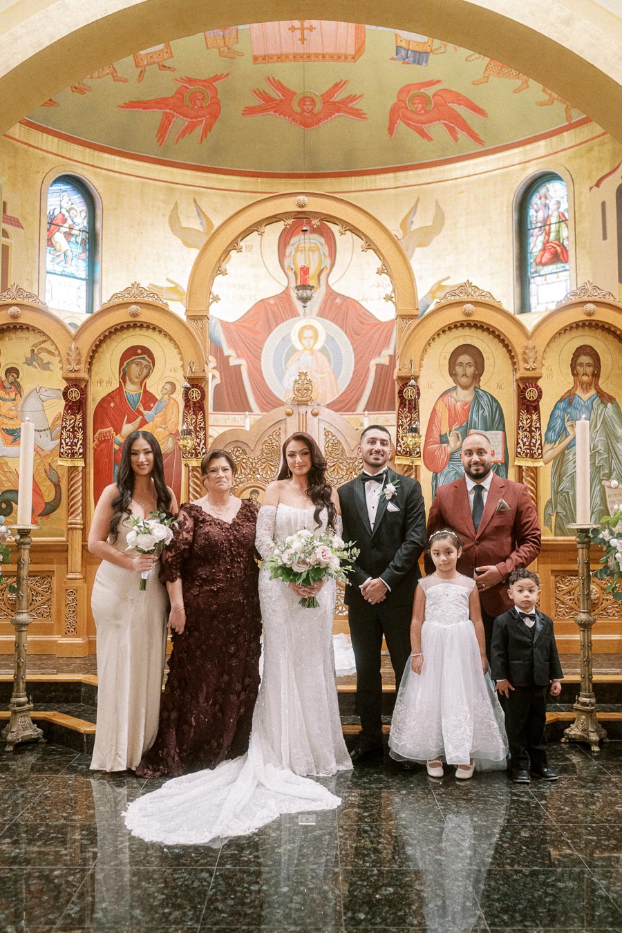 A wedding party poses inside an ornate church, featuring a richly decorated altar with religious iconography. The bride and