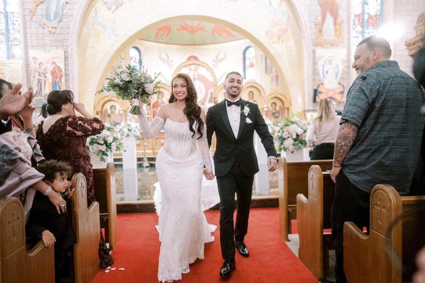Newlywed couple joyfully exiting a beautifully decorated church, with the bride holding a bouquet of flowers and guests