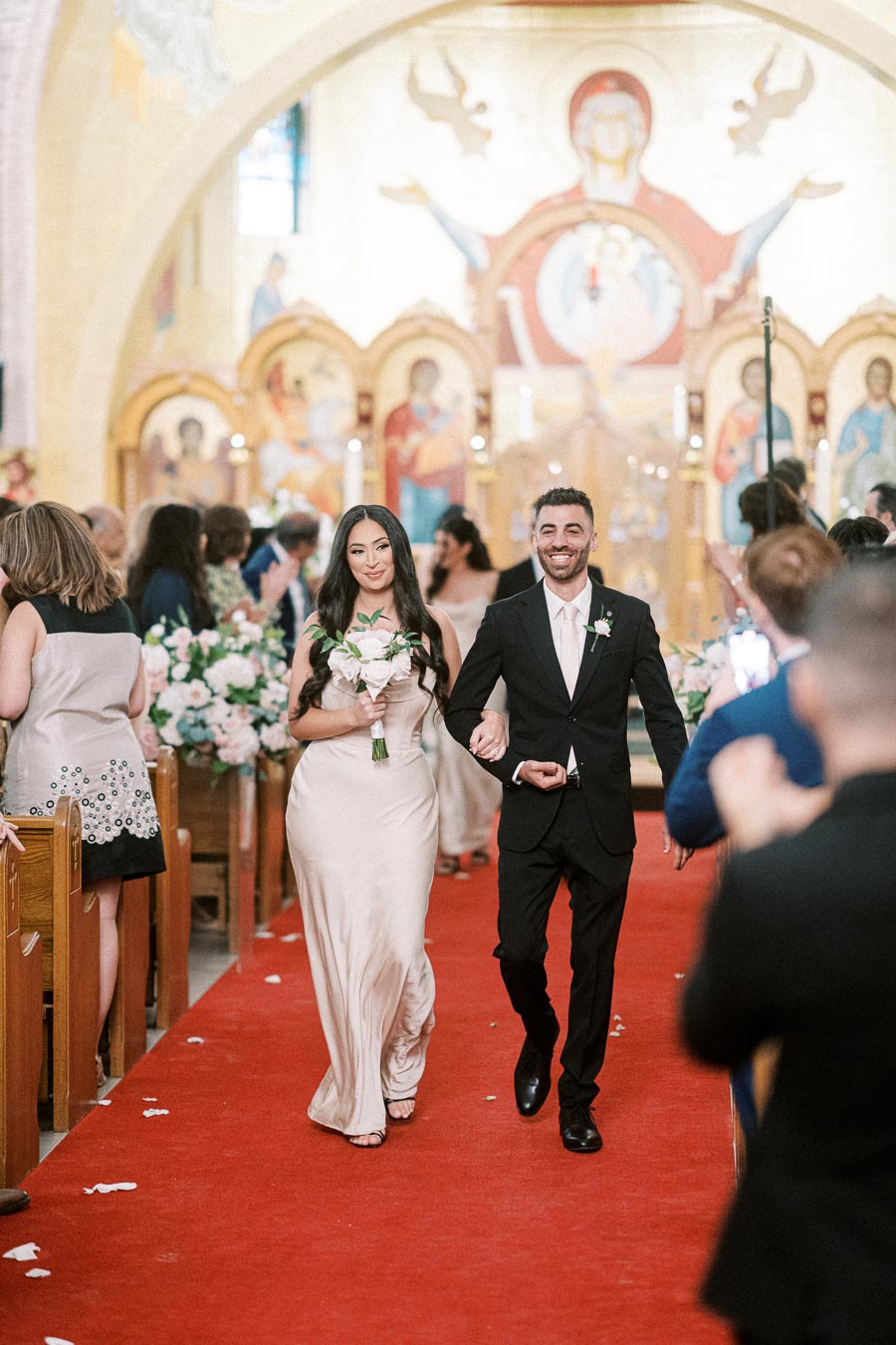 A couple joyfully walking down the aisle in a beautifully decorated church, with the woman holding a bouquet of flowers,