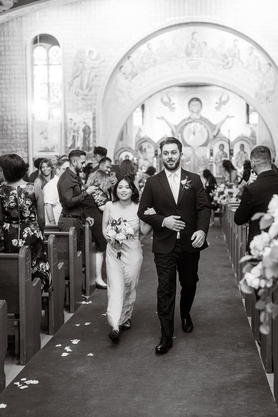 A black and white image of a joyful wedding ceremony in a church, featuring a couple walking arm-in-arm down the aisle,