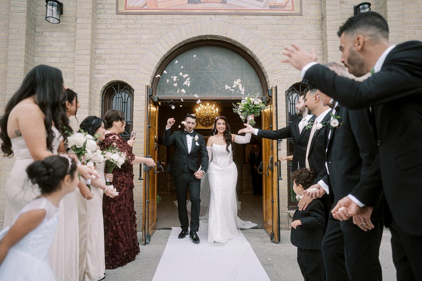 Bride and groom exiting church surrounded by cheering wedding guests, flower petals in the air, and a joyful atmosphere.