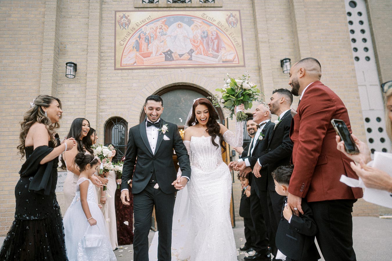 A joyful wedding couple exits a chapel with friends and family celebrating, featuring a bride in a white gown holding a