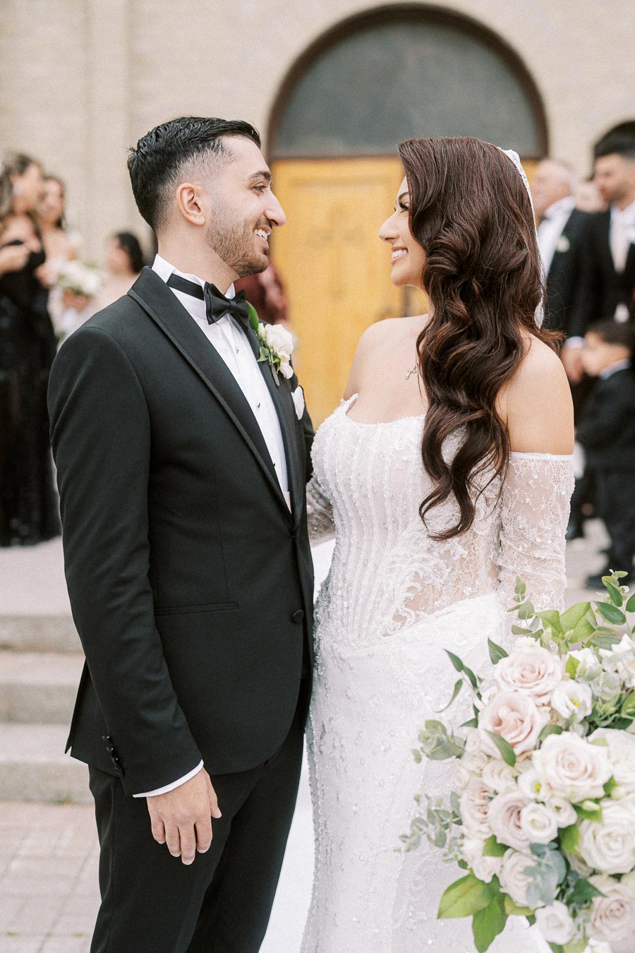 A newlywed couple stands smiling at each other in front of a church entrance, wearing a black tuxedo and a lace wedding