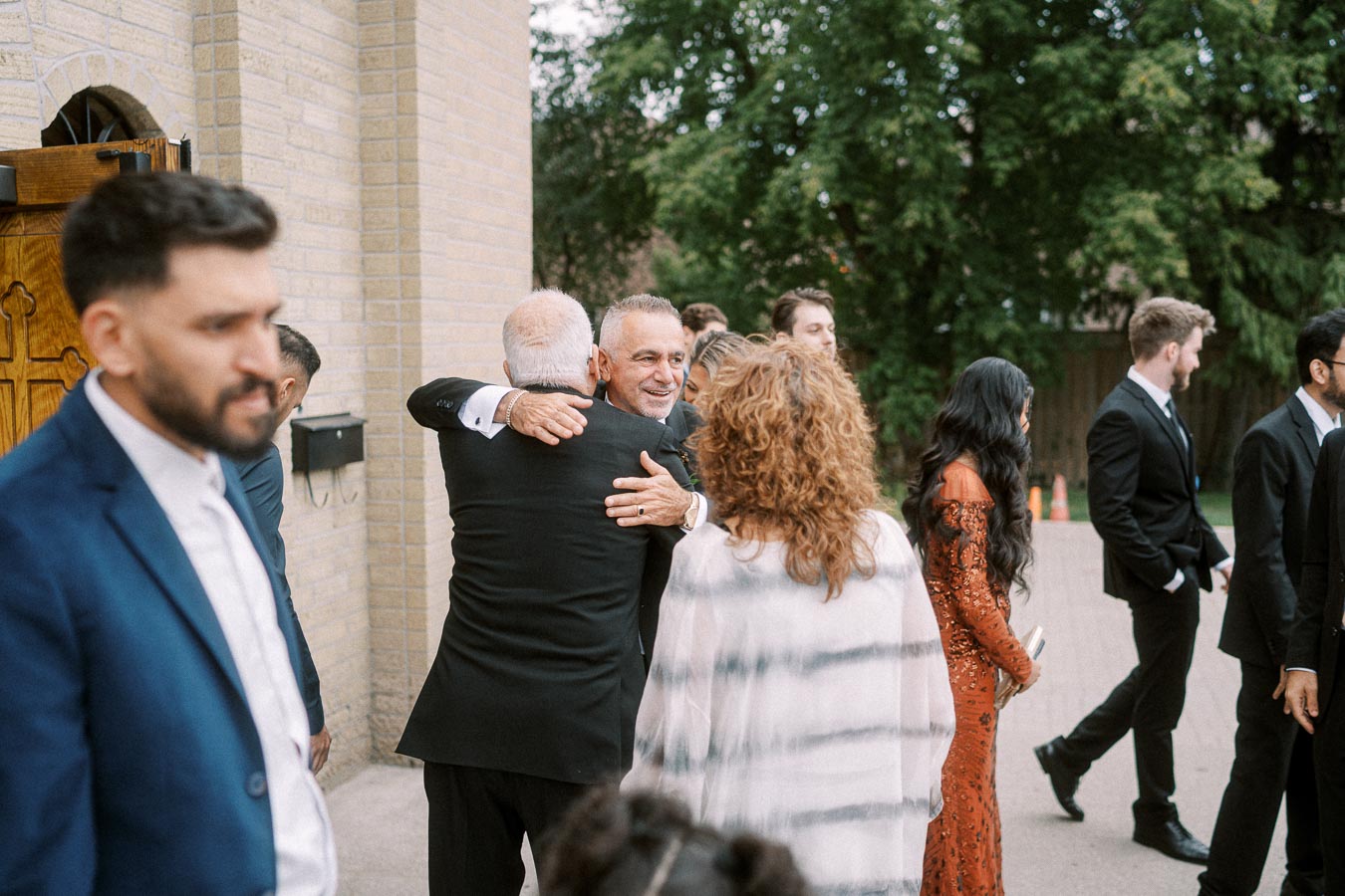 A group of people in formal attire gather outside a brick building, with one man warmly embracing another. Trees can be seen