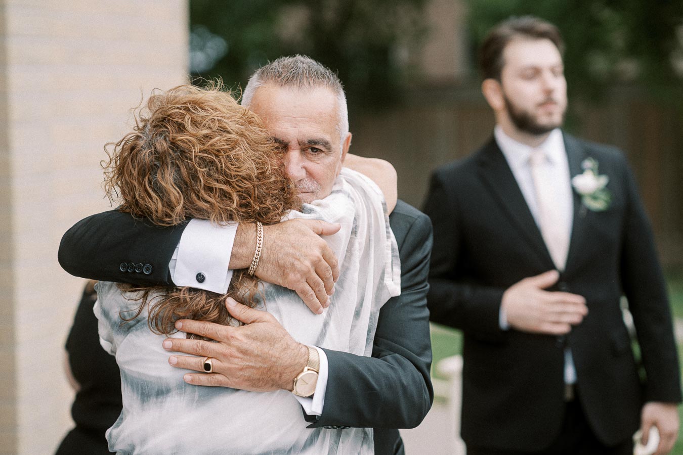 A man in a suit warmly embraces a woman, conveying emotions of support and love, with another man in a suit standing in the