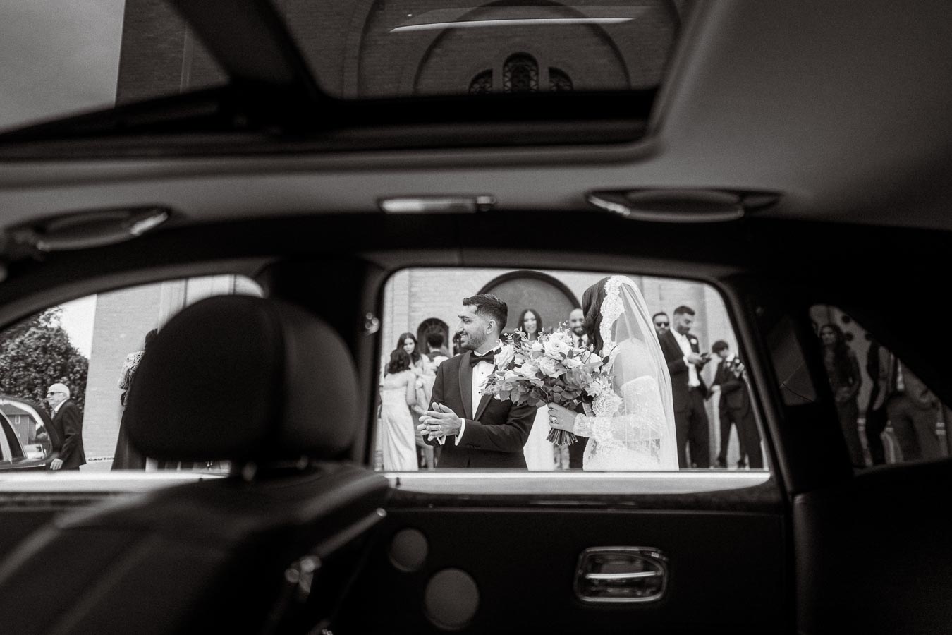 Black and white image of a bride and groom seen from a car window, surrounded by wedding guests outside a building, with the