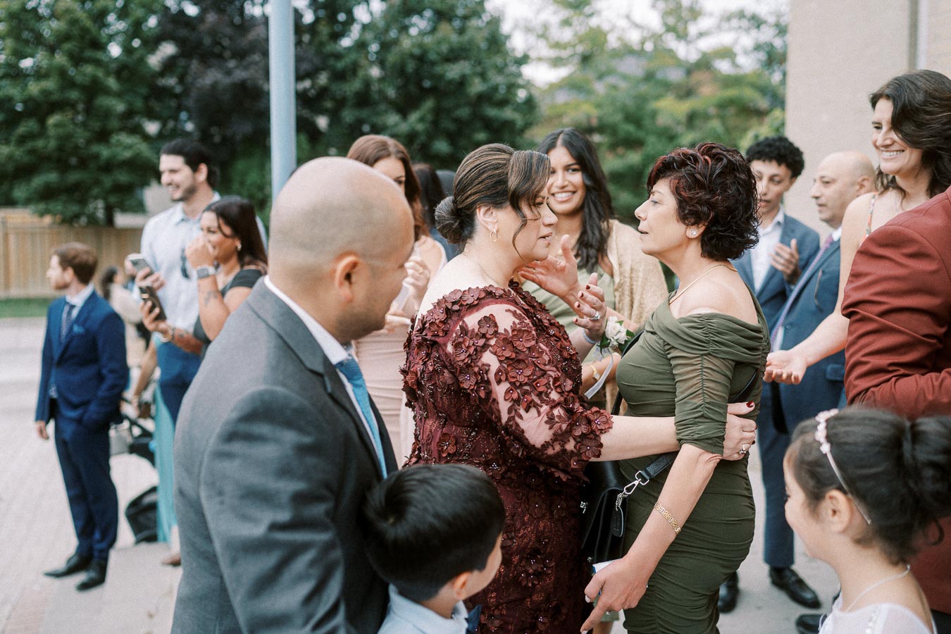 A diverse group of people dressed in formal attire, smiling and interacting at an outdoor social event, with greenery in the
