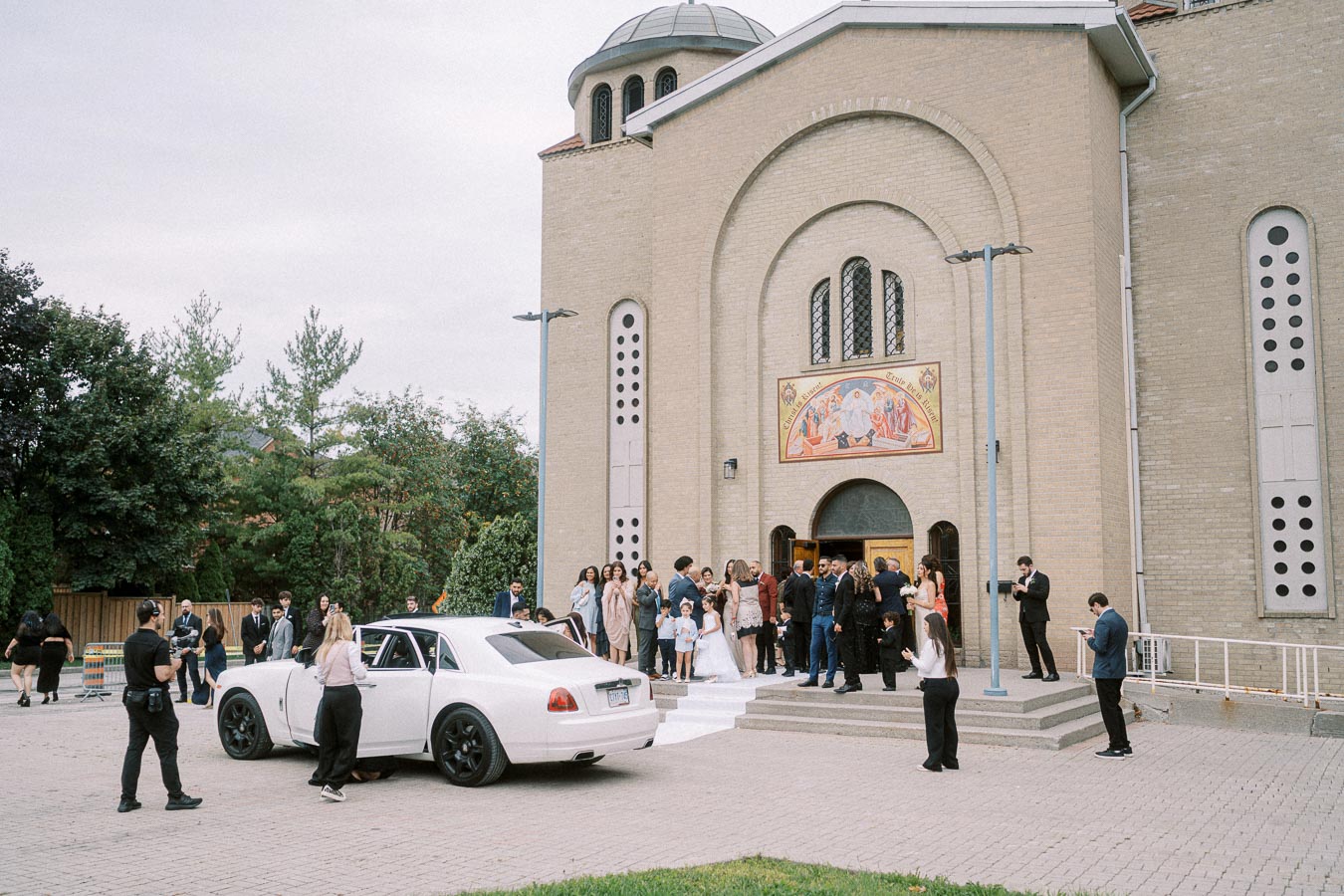 A group of people gathered outside a large brick church, with a white luxury car parked in front.