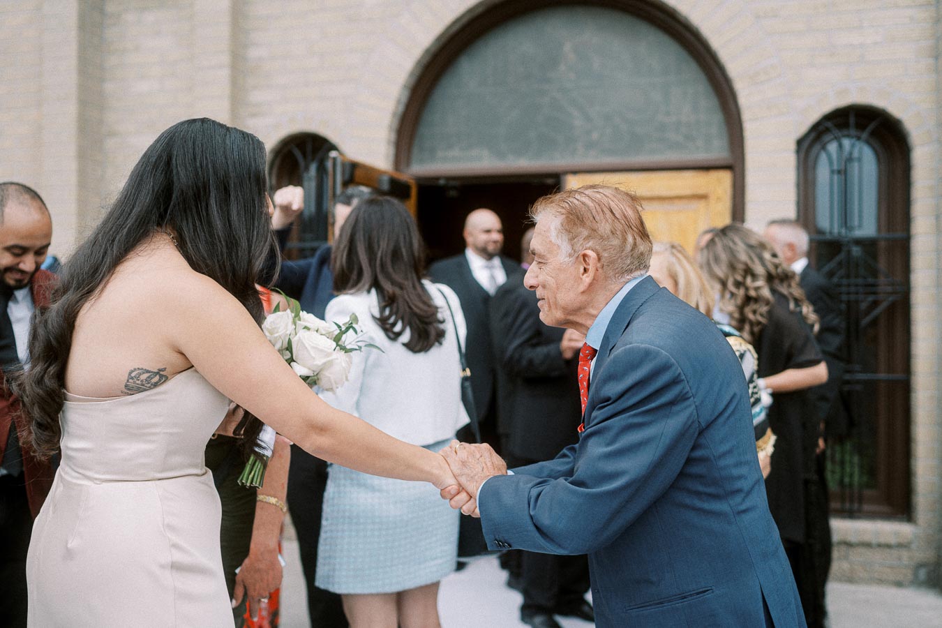 A young woman in a strapless dress shakes hands with an older man in a suit outside a brick building. She holds a bouquet of