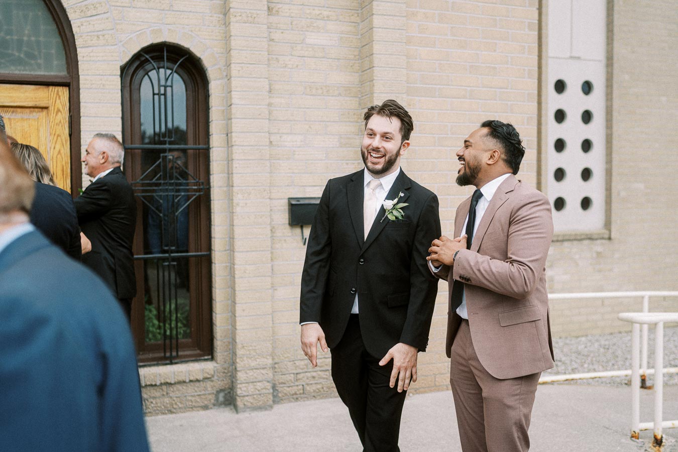 Two men in suits laughing and talking outside a brick building, one in a black suit with a boutonniere and the other in a