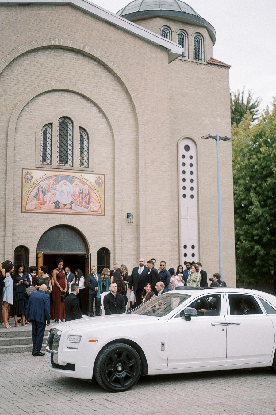 A white Rolls Royce parked outside a stone church with a dome, surrounded by a group of elegantly dressed people attending a