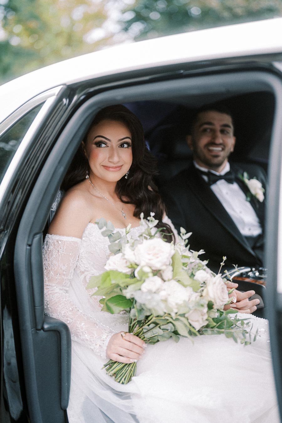 Elegant bride holding a bouquet of flowers, sitting in a car with the groom, both smiling in a candid wedding moment.