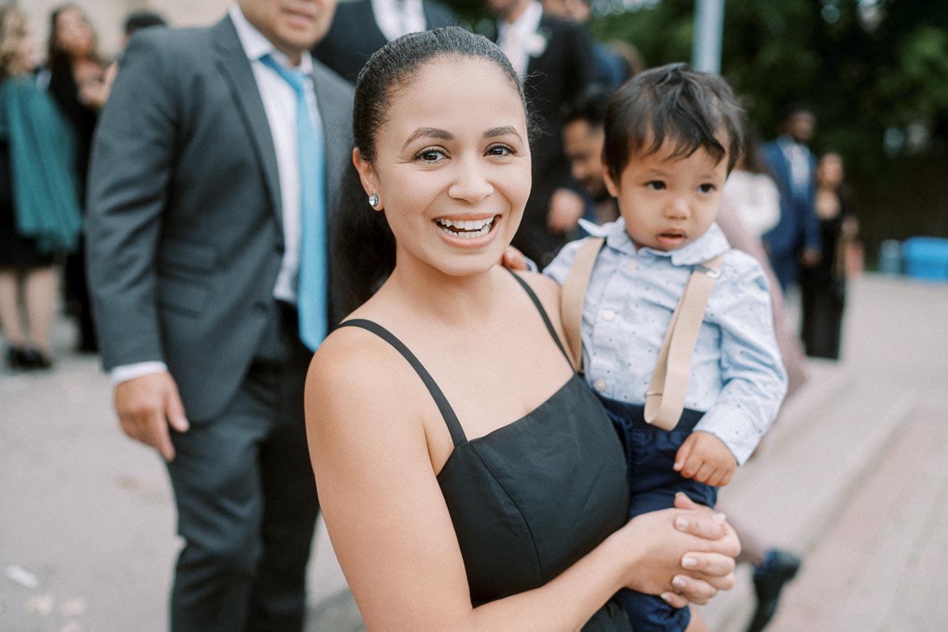 A woman in a black dress smiles while holding a child dressed in a blue shirt and suspenders, with people in formal attire