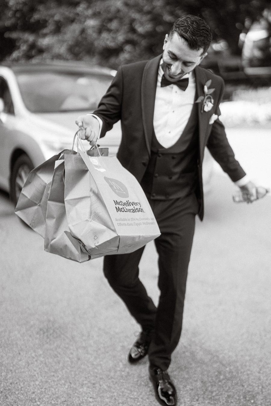 A man in a suit and bow tie joyfully carries multiple takeaway bags labeled McDelivery, walking past a car on a sunny day.