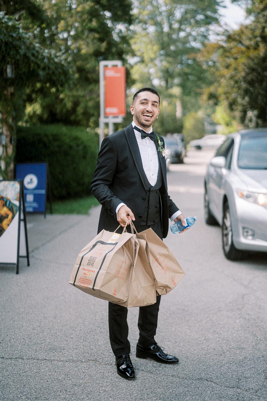 A smiling man in a black tuxedo carries grocery bags and a water bottle on a sunny day, standing on a street with a parked