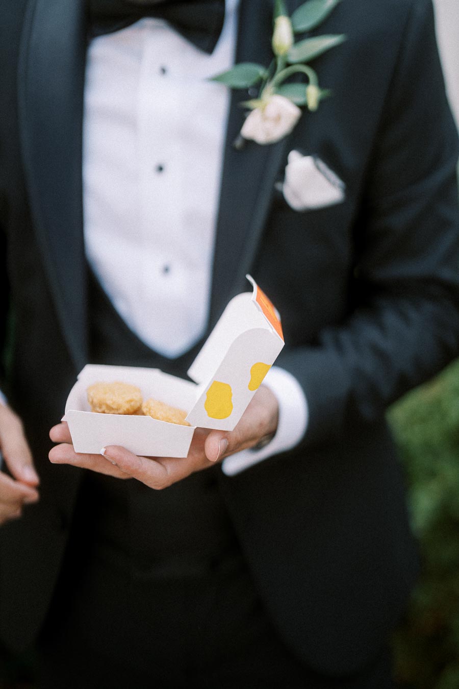 A groom in a tuxedo holds a box of chicken nuggets, highlighting a casual moment at a wedding.
