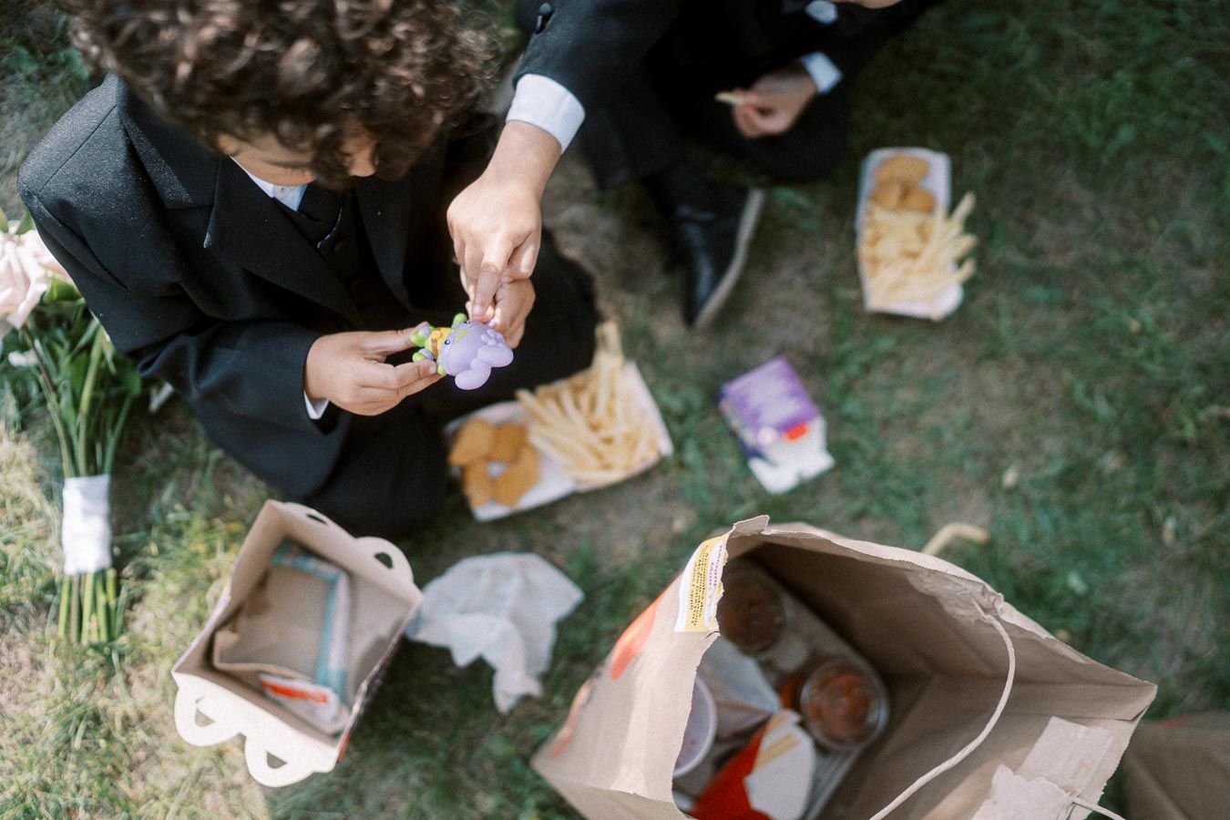 Top view of children in formal attire enjoying a picnic with fast food on a grassy field, including fries and chicken