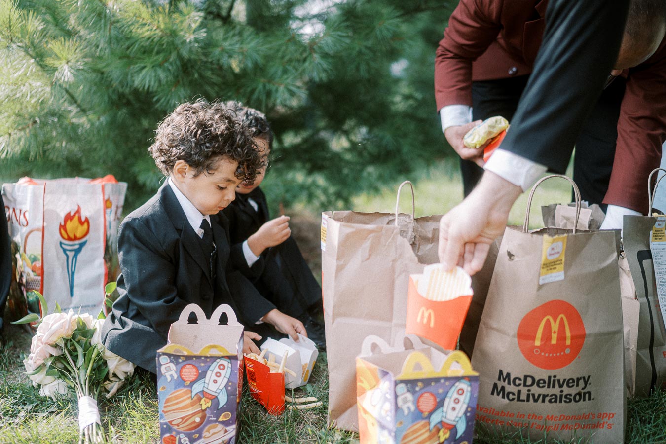 A young child in a suit enjoys a McDonald's Happy Meal, sitting on grass with a bouquet beside him, surrounded by takeaway