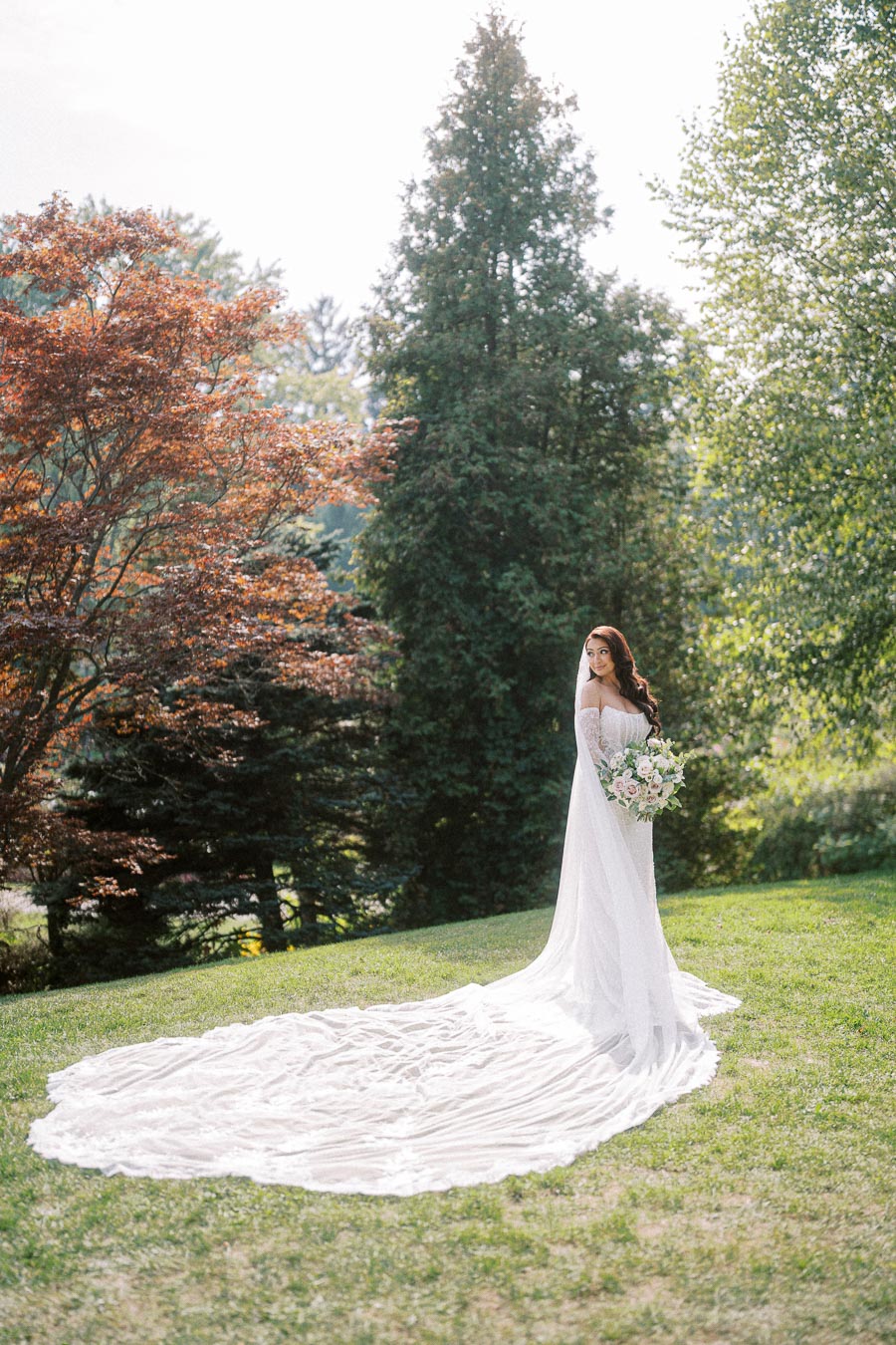 Bride in elegant wedding gown with long train, holding a bouquet, standing on lush green grass in a tranquil garden setting