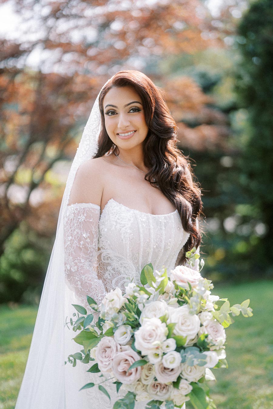Bride in a lace wedding dress holding a bouquet of white and pink roses, standing outdoors with green foliage and soft