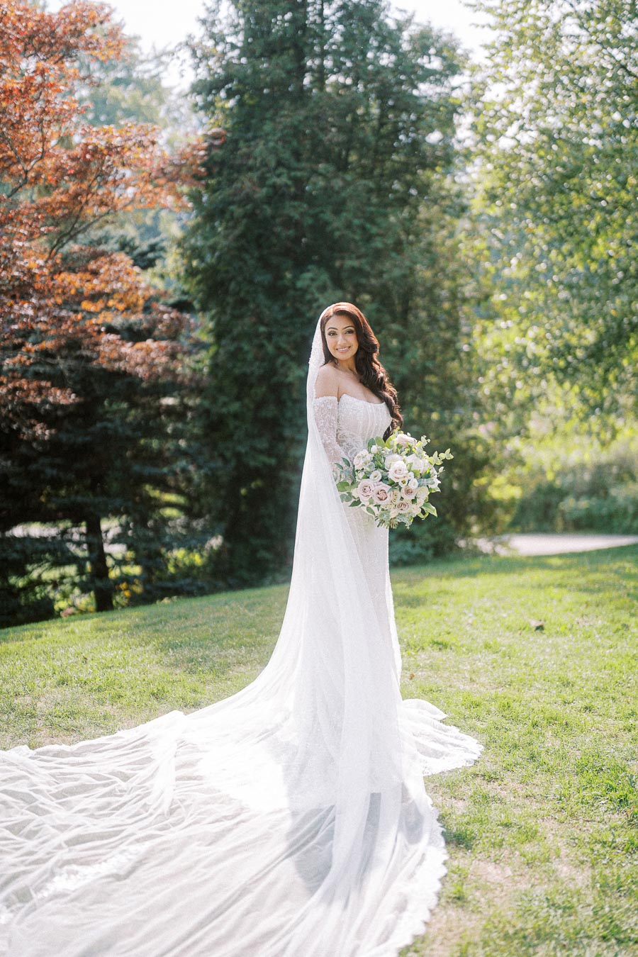 A bride in an elegant long-sleeved lace wedding dress with a flowing train and veil, holding a bouquet of pastel flowers in