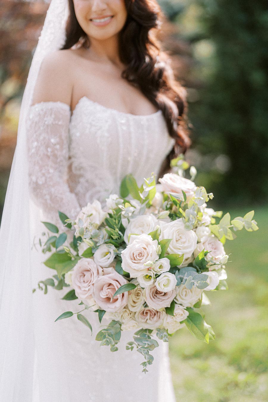 A bride in an elegant lace wedding dress holds a lush bouquet of pale pink and white roses with greenery, set against a