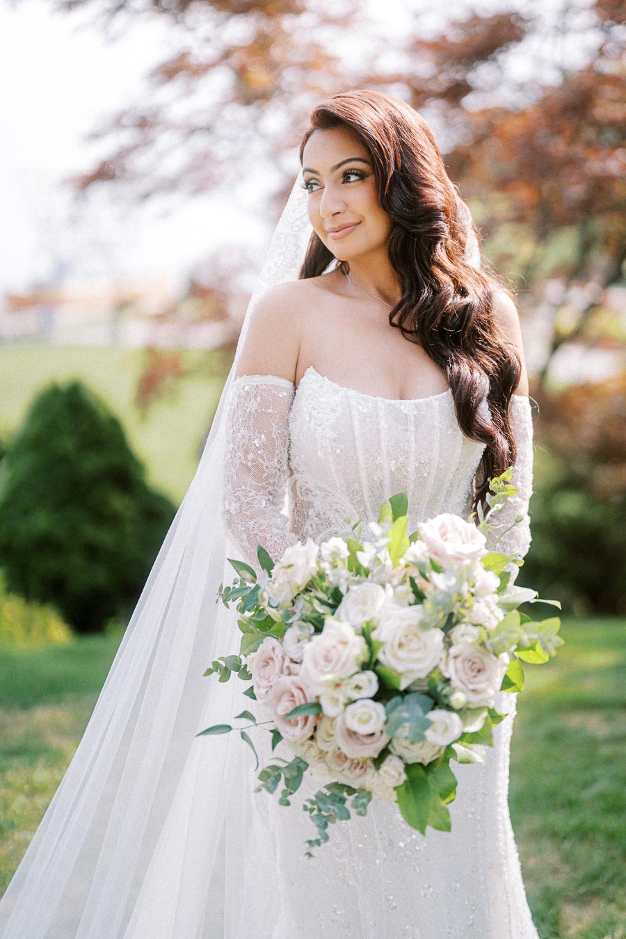 A bride in an elegant lace wedding dress holding a bouquet of pale pink and white roses, standing outdoors with a backdrop