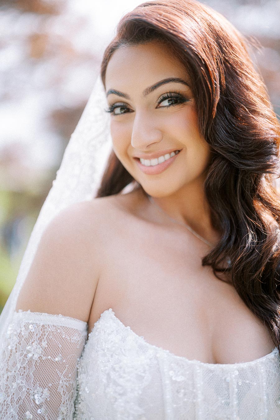 Smiling bride in lace wedding dress with long dark hair and veil, outdoor portrait.