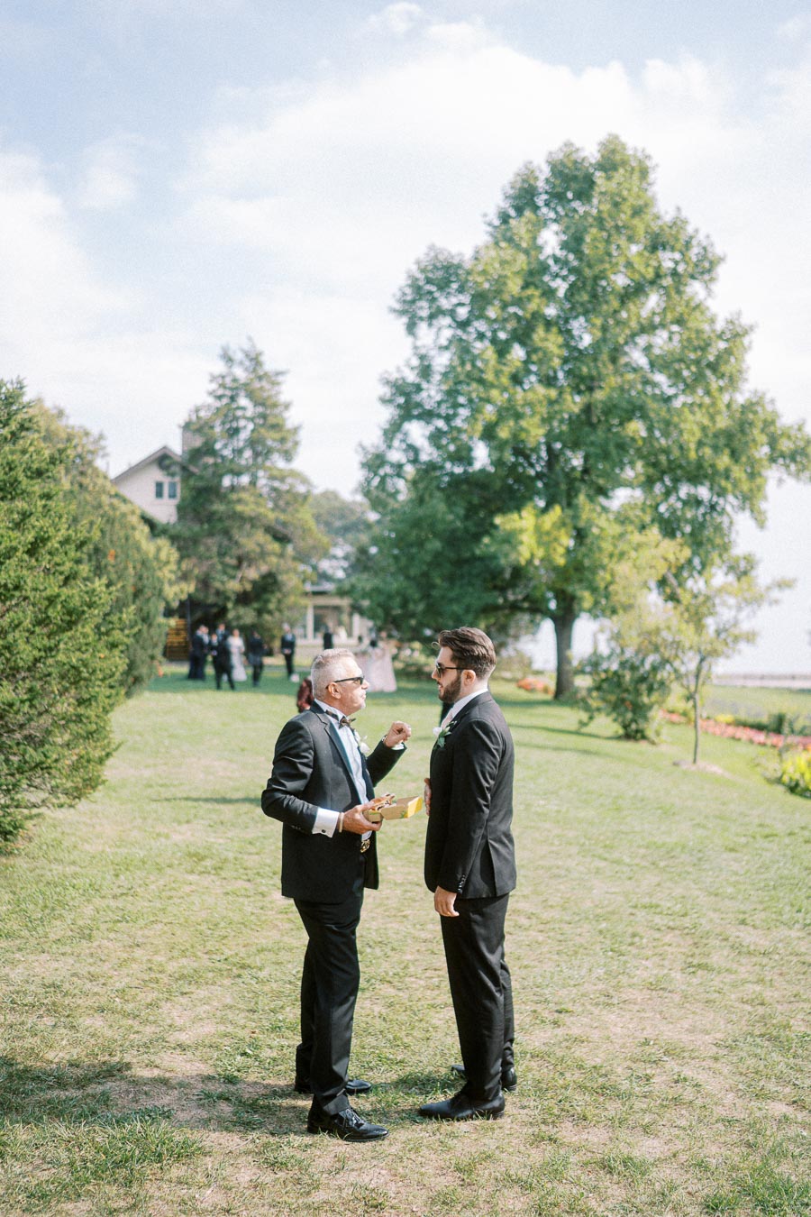 Two men in formal suits having a conversation outdoors on a sunny day, surrounded by greenery and trees, with a house