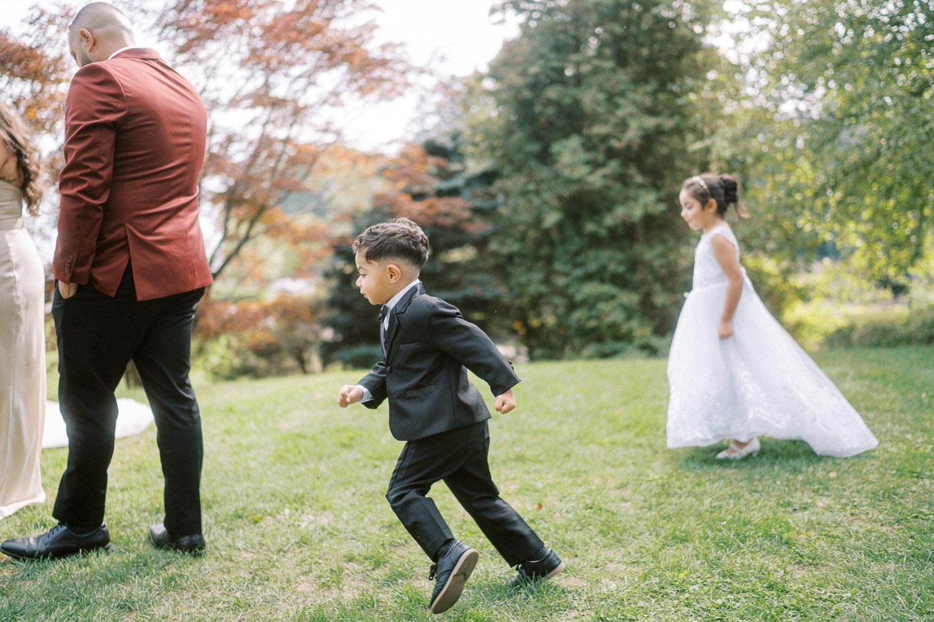 A young boy in a black suit playfully runs across a grassy area, accompanied by a young girl in a white dress. They are part