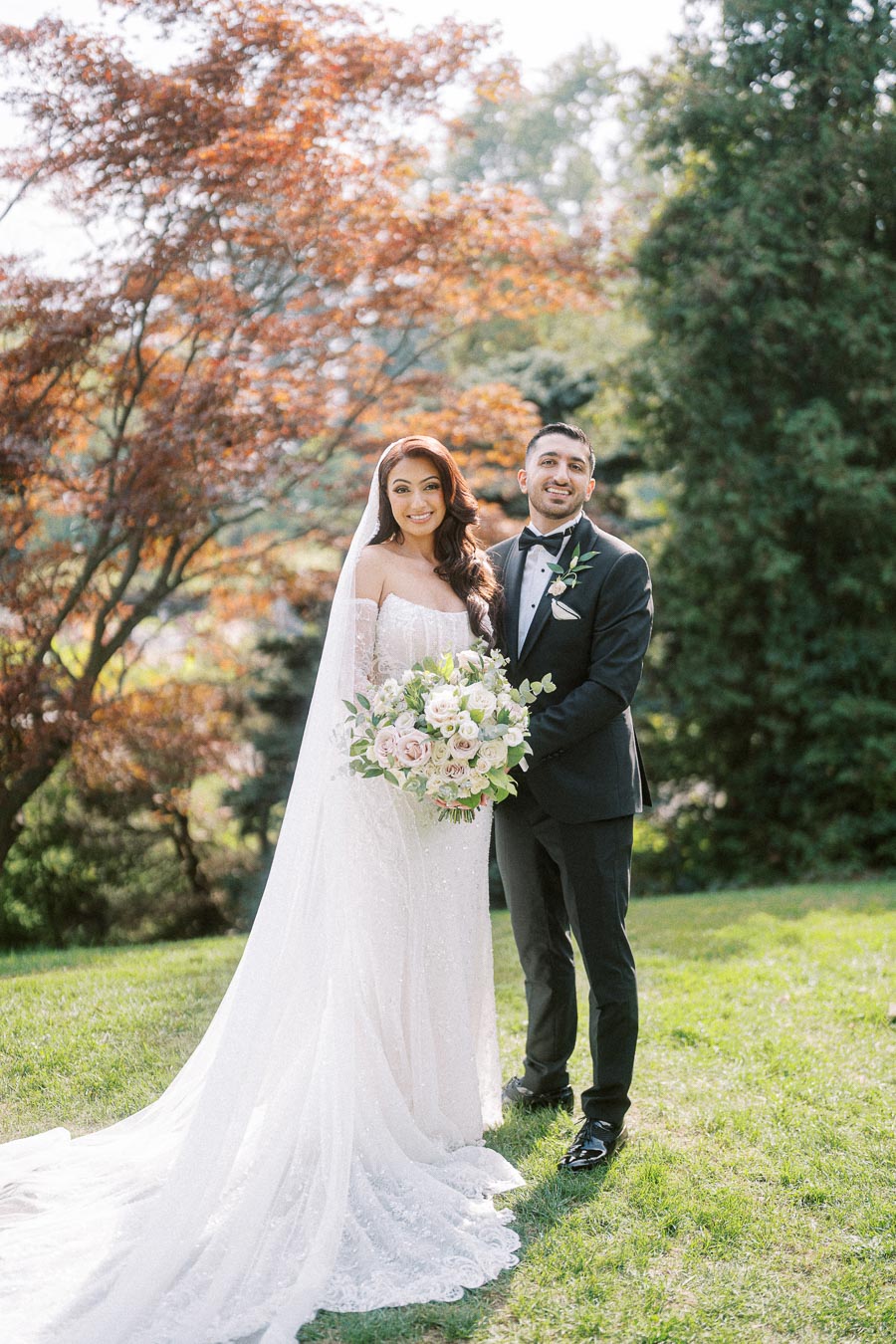 Elegant bride in a lace wedding dress holding a bouquet, standing with the groom in a black tuxedo in a sunlit garden with