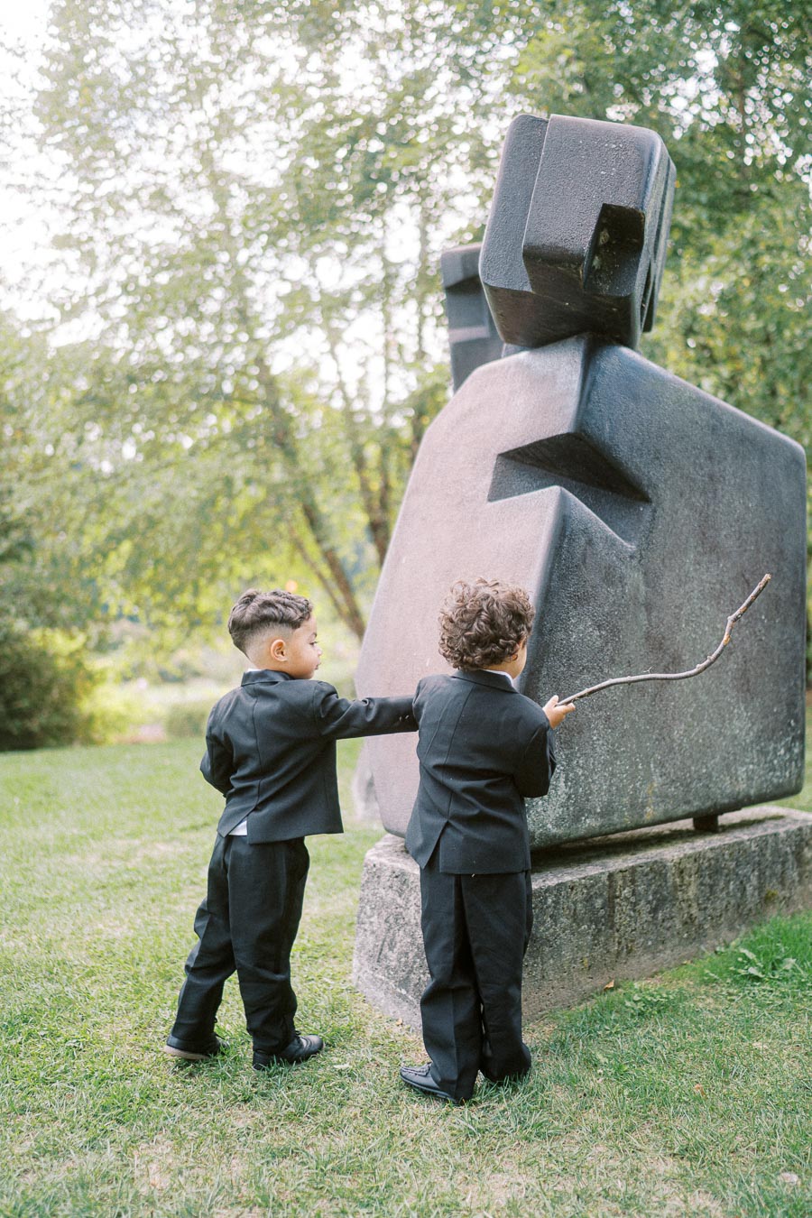 Two young boys in black suits curiously interact with a large abstract stone sculpture in a lush green park. One boy points