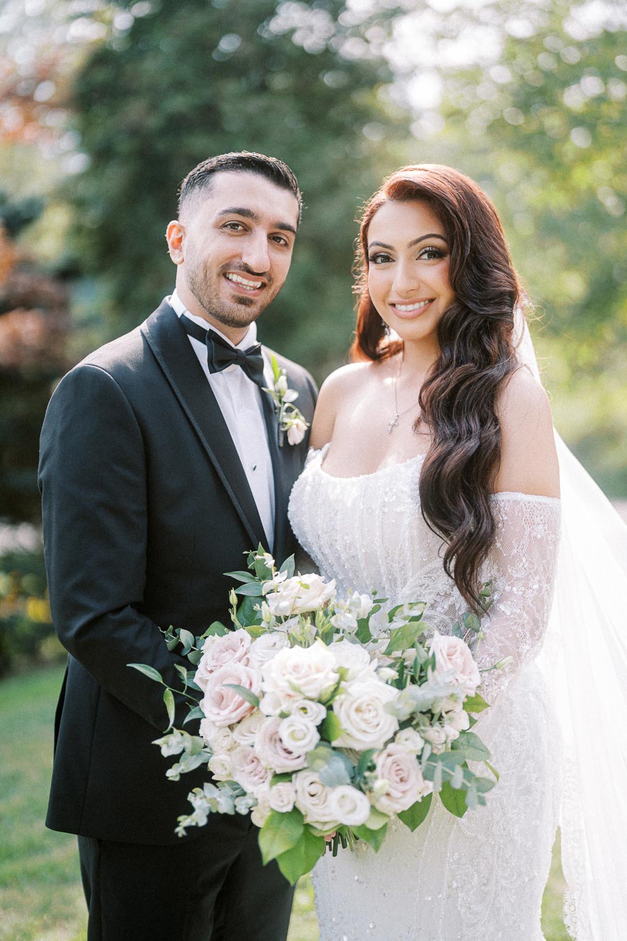 Happy couple in formal attire on wedding day, bride holding bouquet of roses, outdoor setting with soft lighting.