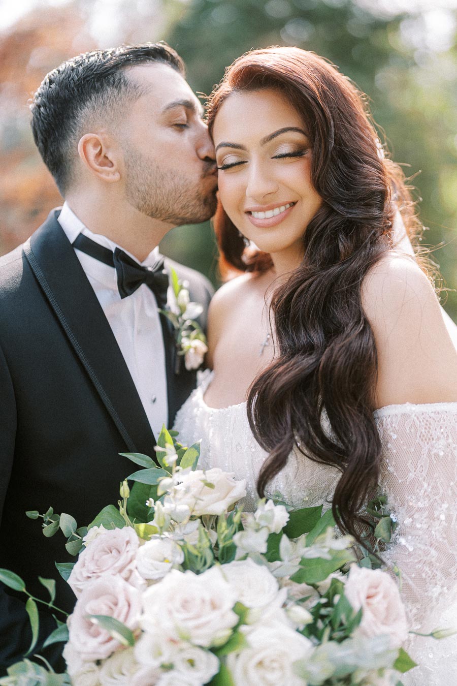 A happy couple on their wedding day, with the groom in a black tuxedo gently kissing the bride, who is holding a bouquet of
