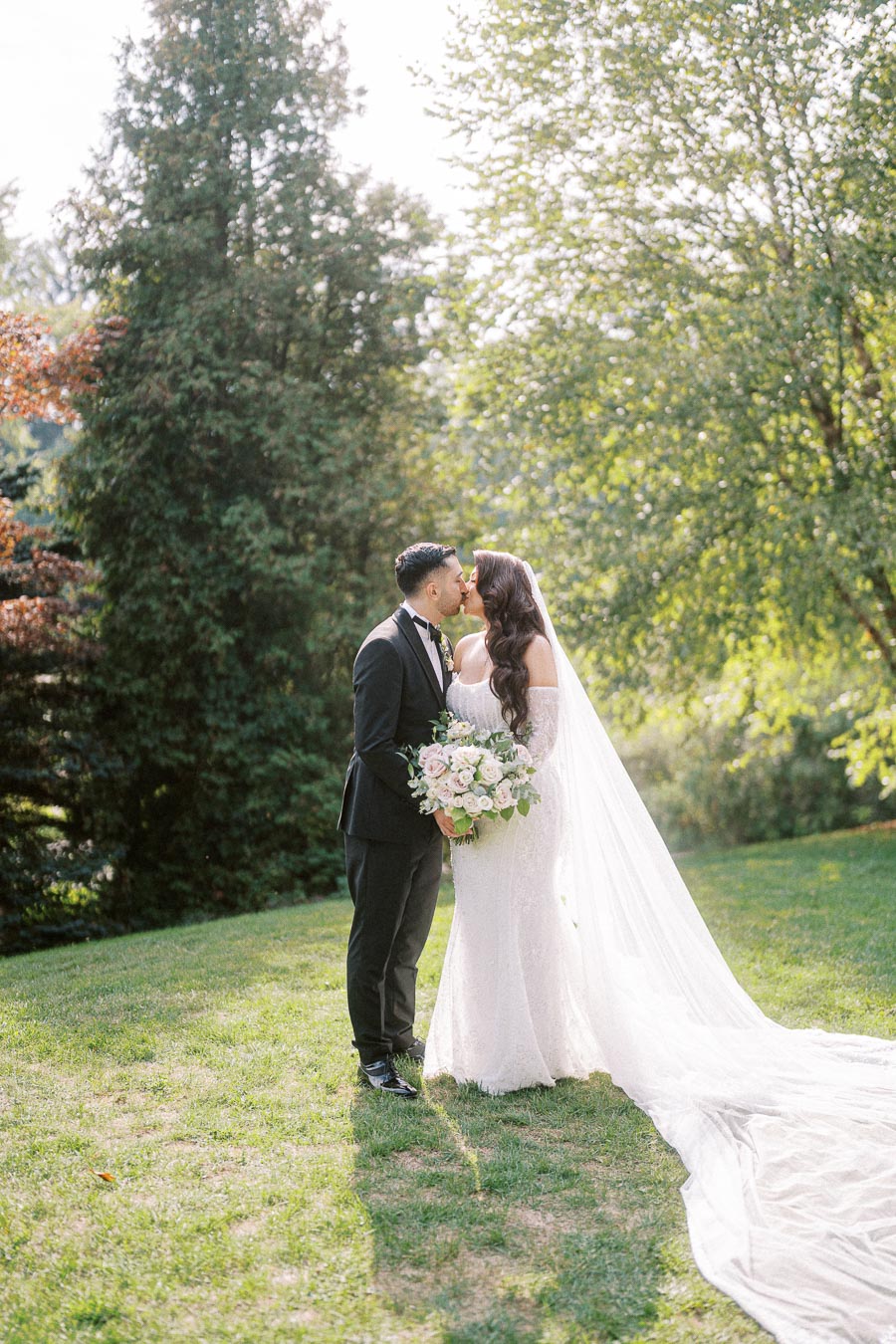 A bride and groom share a romantic kiss in a sunlit garden, with the bride wearing a flowing white gown and veil and holding