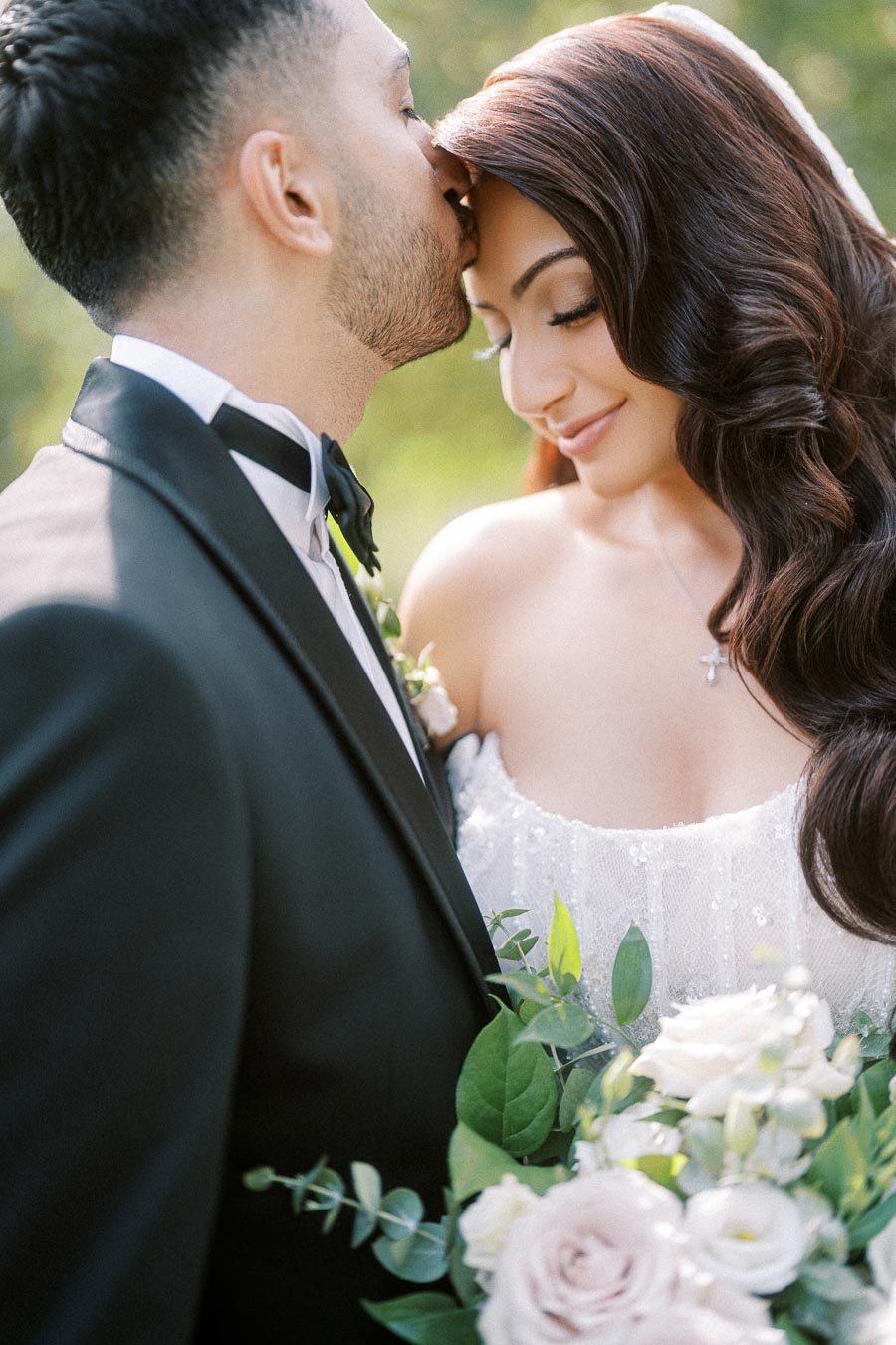 Elegant wedding couple, groom in black tuxedo kissing bride's forehead, holding bouquet of white and blush roses outdoors.