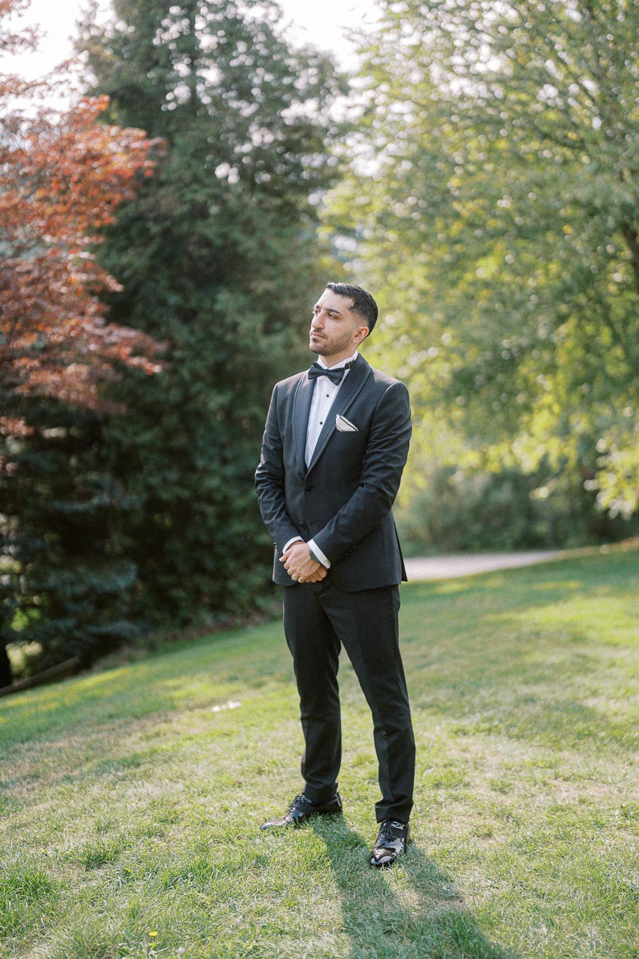 Man in a tuxedo standing outdoors on a sunny day with lush greenery in the background, exuding elegance and style.