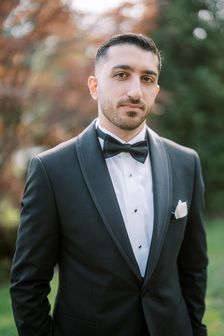 A man in a formal black tuxedo with a bow tie stands outdoors, looking at the camera with a blurred natural background.