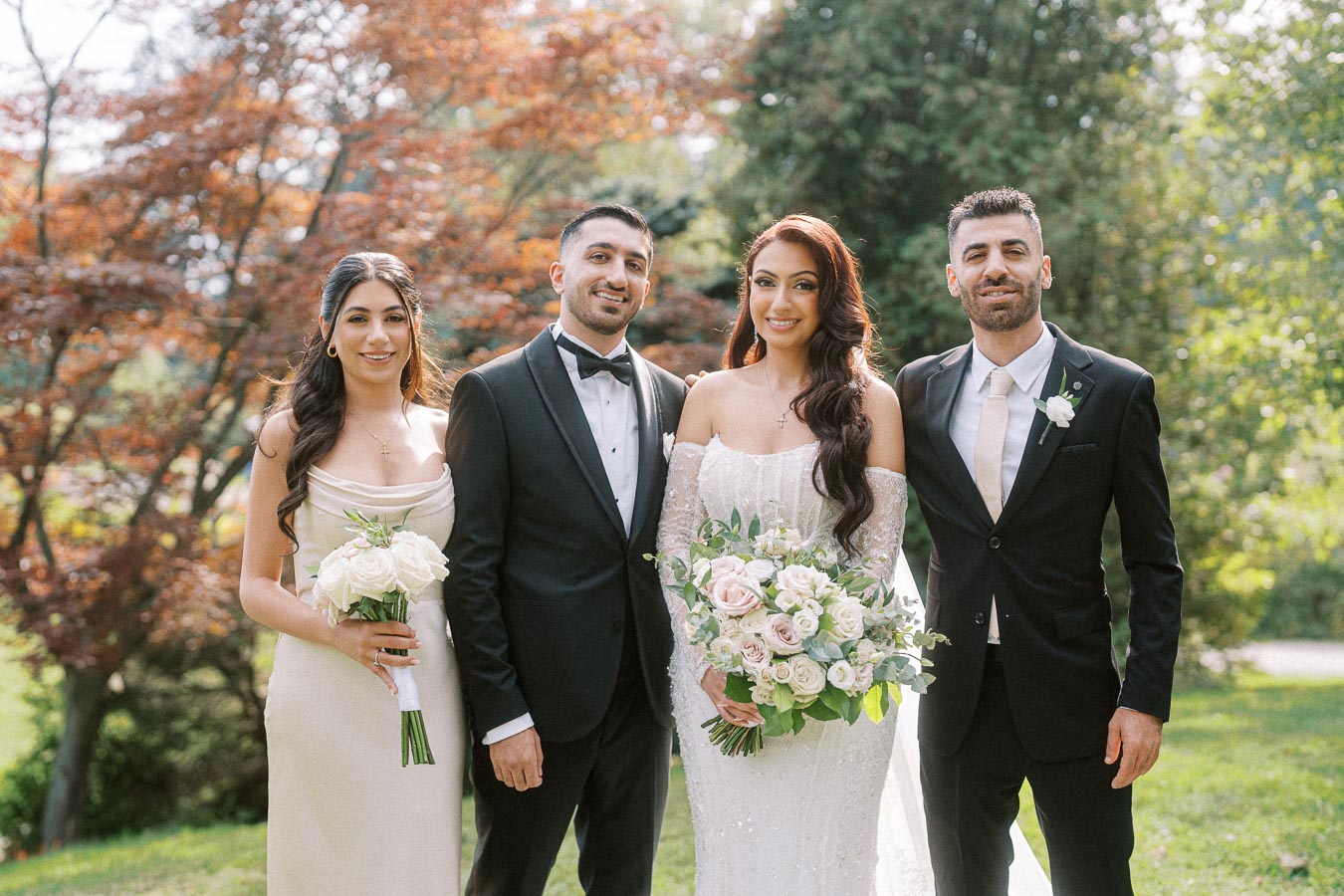 A happy wedding couple poses with two guests in elegant attire in a lush garden setting, with the bride holding a vibrant