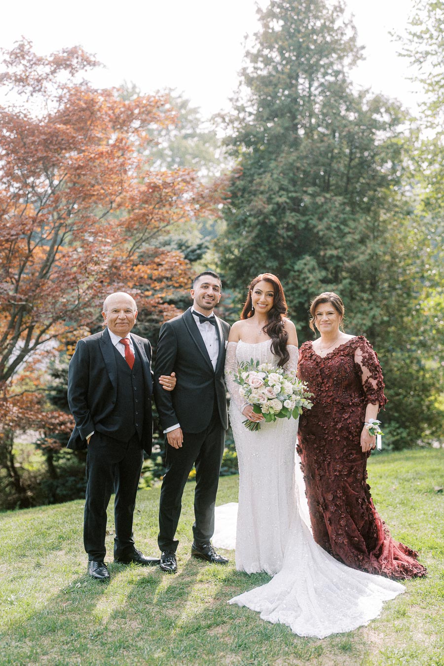 Wedding couple with parents posing outdoors on a sunny day, with autumn trees in the background.