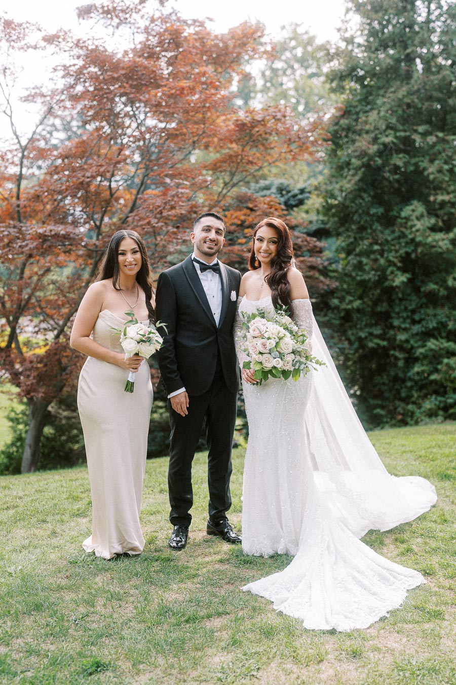 Three people pose outdoors; a woman in a strapless gown holding a bouquet, a man in a tuxedo, and a bride in a white wedding