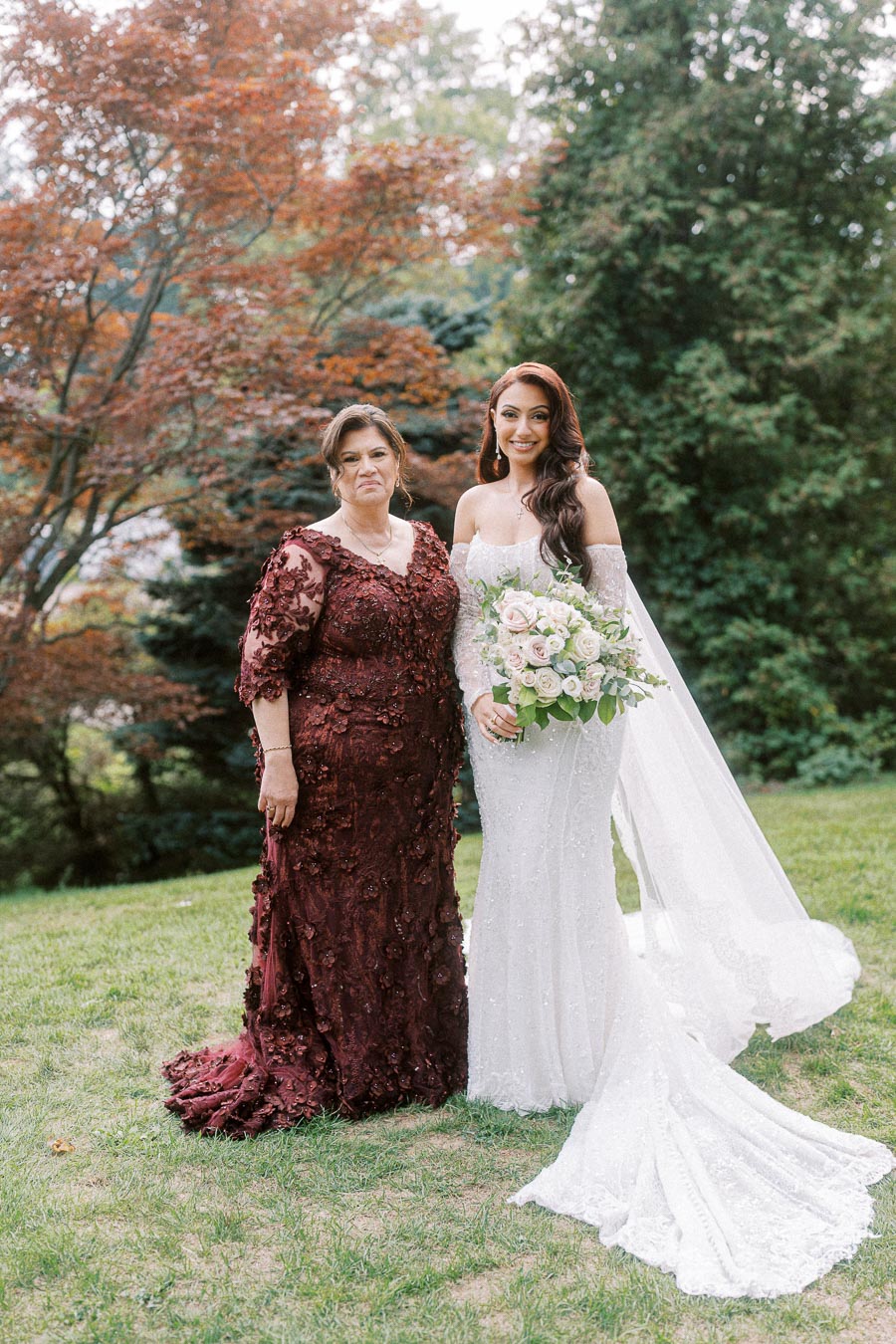 Mother and bride posing together outdoors in elegant dresses; the bride wears a flowing white gown and holds a bouquet,