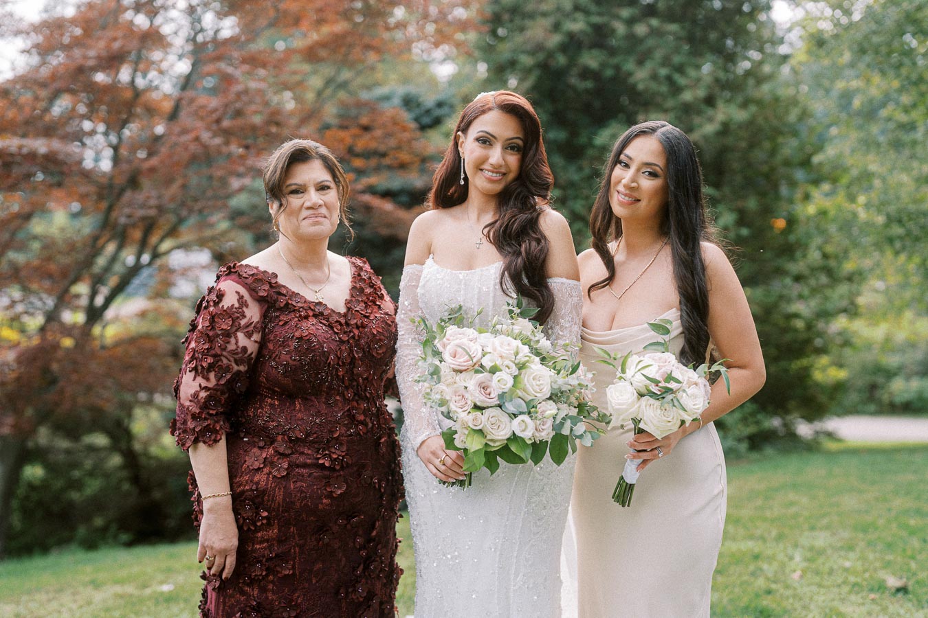 Three women pose together outdoors, with one in a bridal gown holding a bouquet of roses, flanked by two women in formal